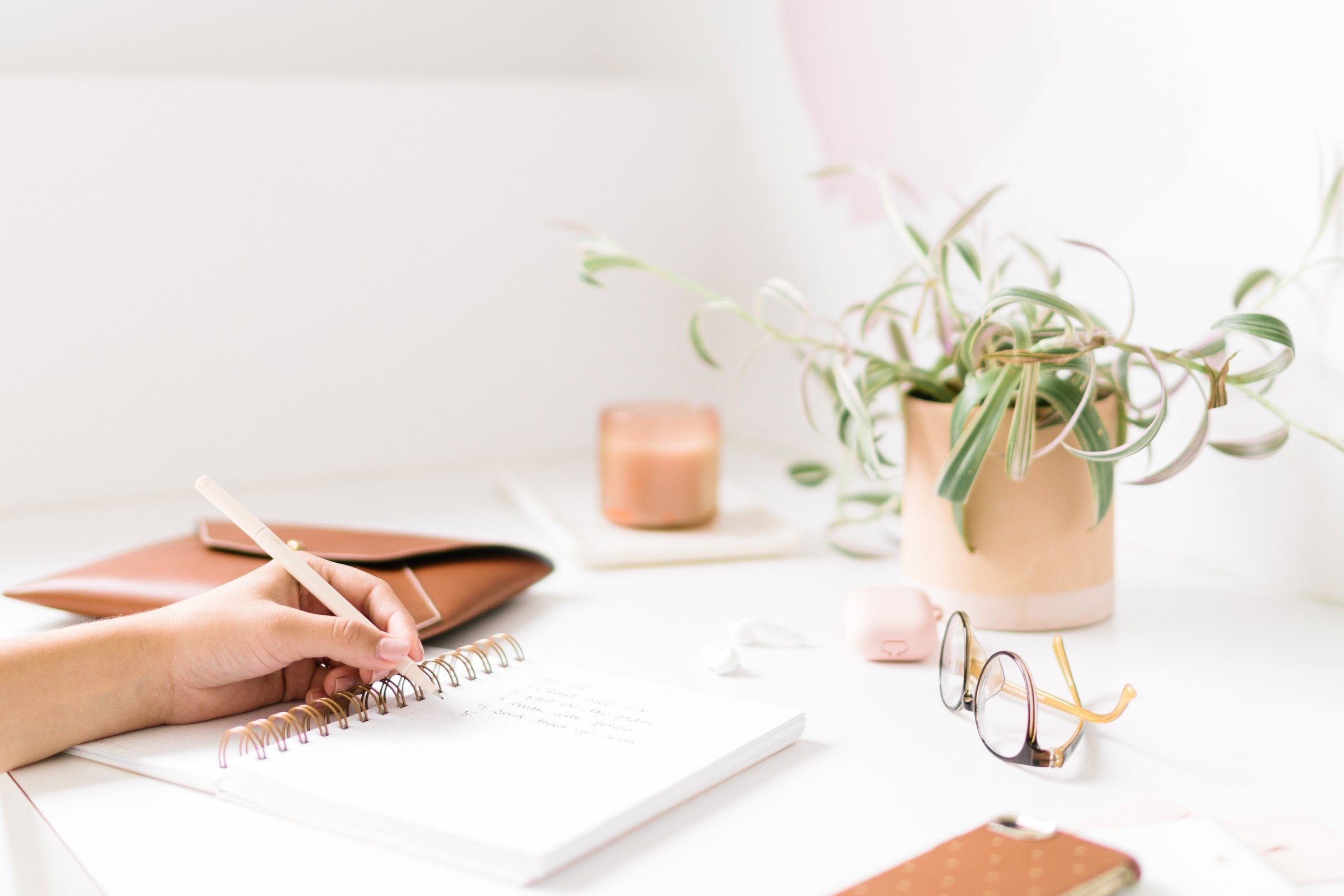 Person writing in a notebook on a desk with plants, glasses, a phone and other clutter